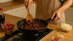 Seorang wanita sedang memasak olahan daging sapi di dapur modern, menggunakan wajan anti lengket di atas kompor gas. Di sekelilingnya terdapat bahan-bahan segar seperti bawang bombai, bawang putih, cabai merah, dan potongan daging sapi mentah yang siap diolah.